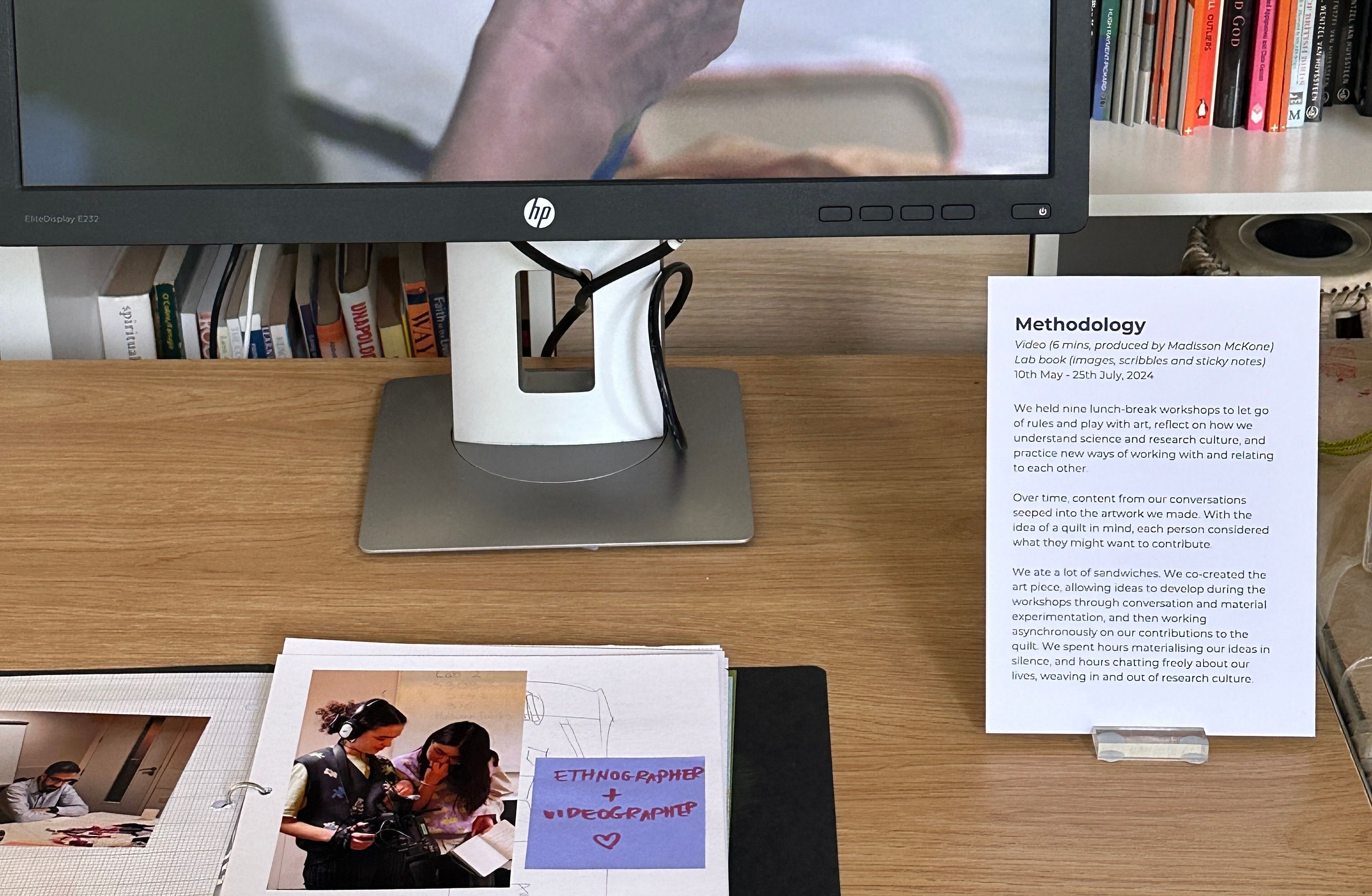 A computer screen and notebook. The notebook has a photo of two people and a note saying ethnographer + videographer. An A4 sheet reads 