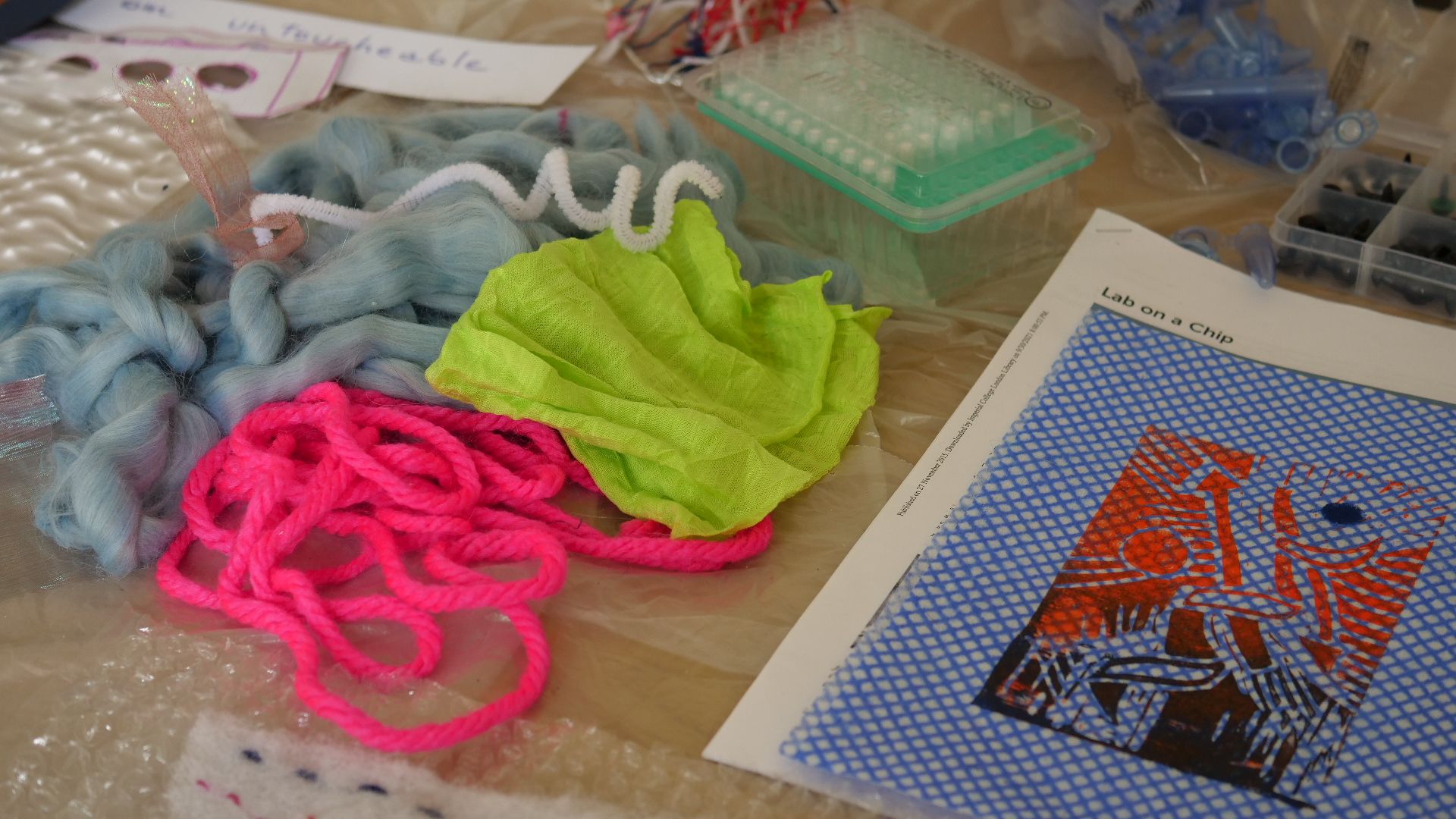 Several art materials (e.g. wool) and laboratory materials on a table; a piece of paper reads 